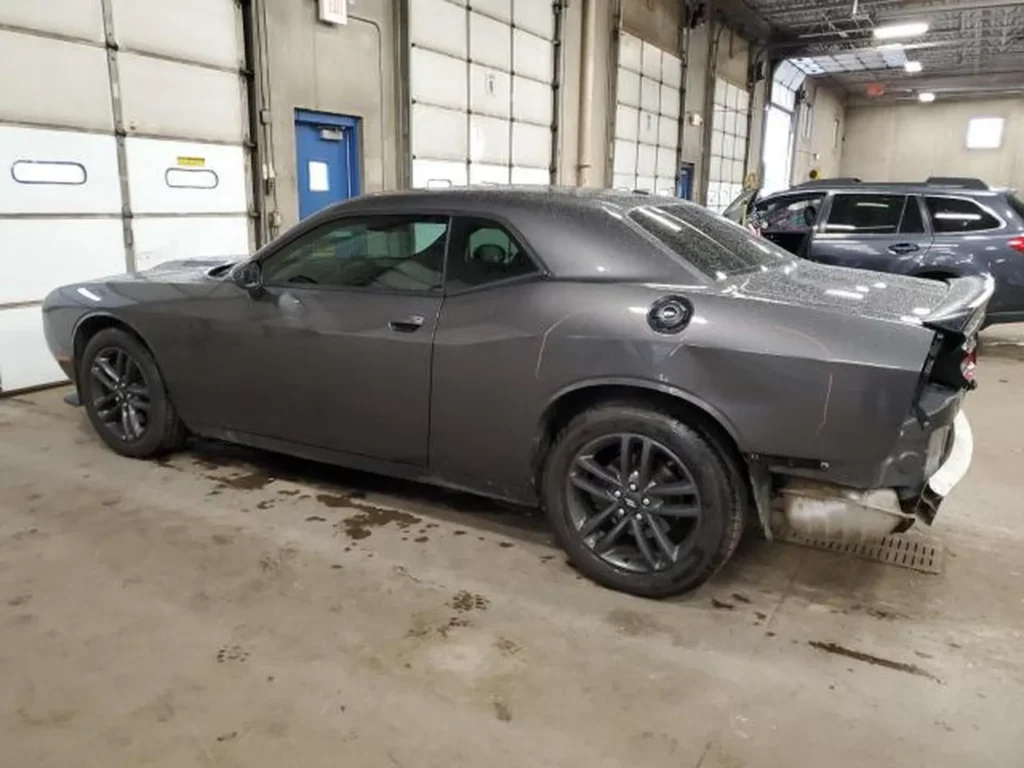 Szary Dodge Challenger coupe z lewej tylnej strony, widoczne uszkodzenie zderzaka w hali warsztatowej.