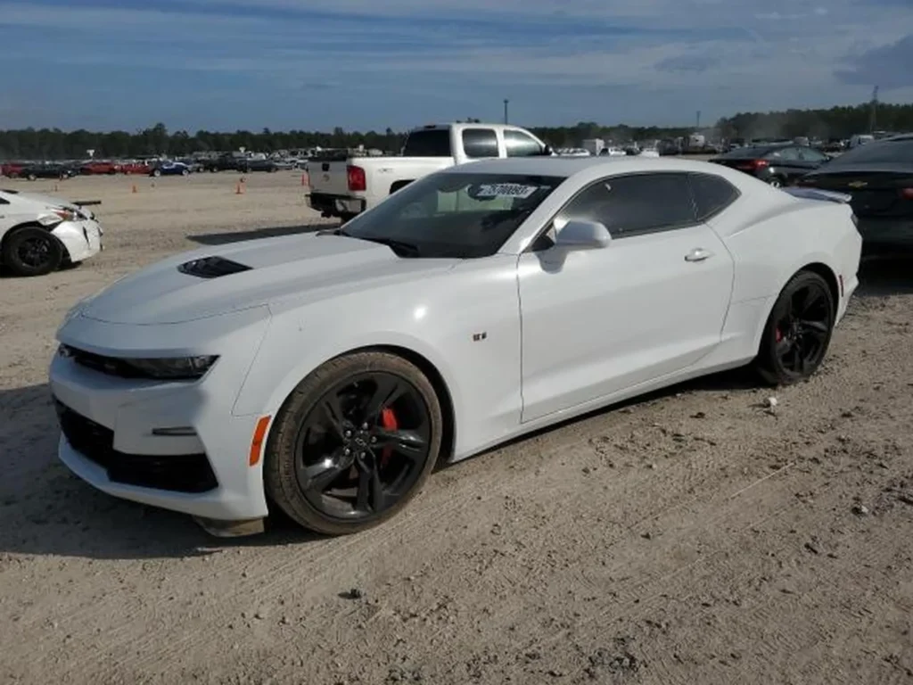 Biały Chevrolet Camaro coupe z przodu z lewej, czarne felgi i czerwone zaciski, plac aukcyjny.