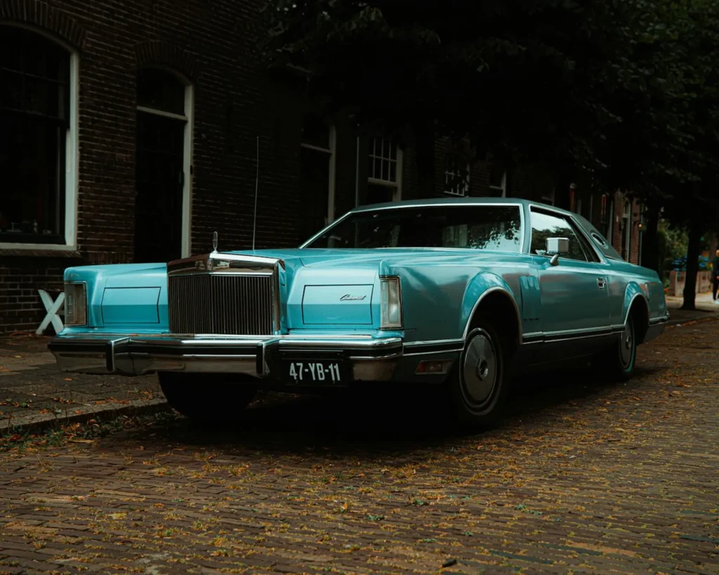 Turkusowy Lincoln Continental Mark V z lat 70. zaparkowany na brukowanej ulicy przy ceglanej kamienicy