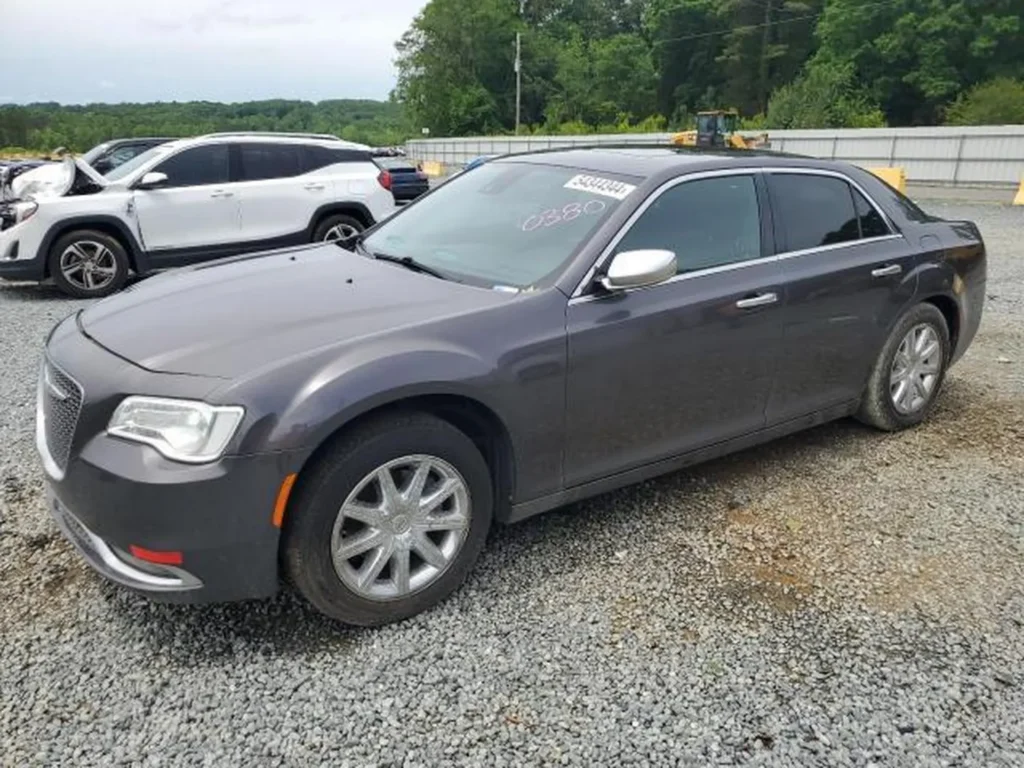 Szary Chrysler 300C (2015) sfotografowany z przodu i lewego boku; auto stoi na żwirowym parkingu obok białego SUV-a.