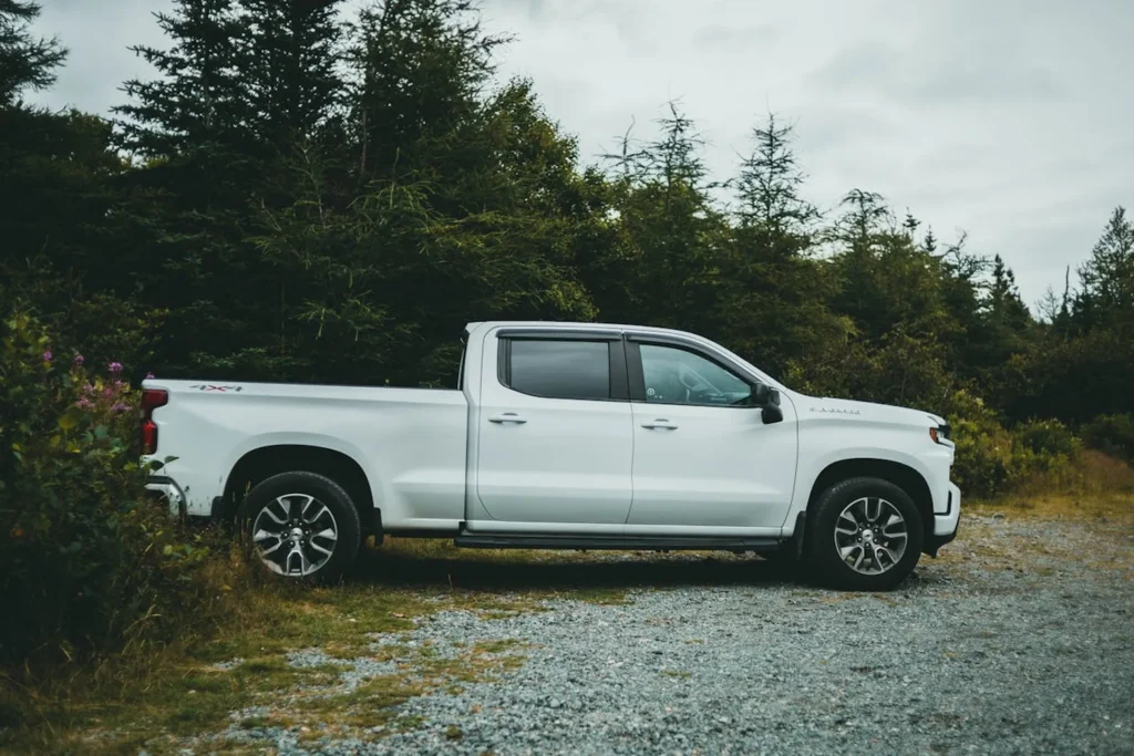 Na zdjęciu widzimy biały pickup Chevrolet Silverado zaparkowany na żwirowej drodze obok gęstego lasu. Samochód ma podwójną kabinę i duże, terenowe opony, co sugeruje, że jest gotowy do jazdy w trudniejszych warunkach terenowych. W tle widoczne są wysokie drzewa iglaste, co nadaje scenerii spokojny, naturalny charakter.