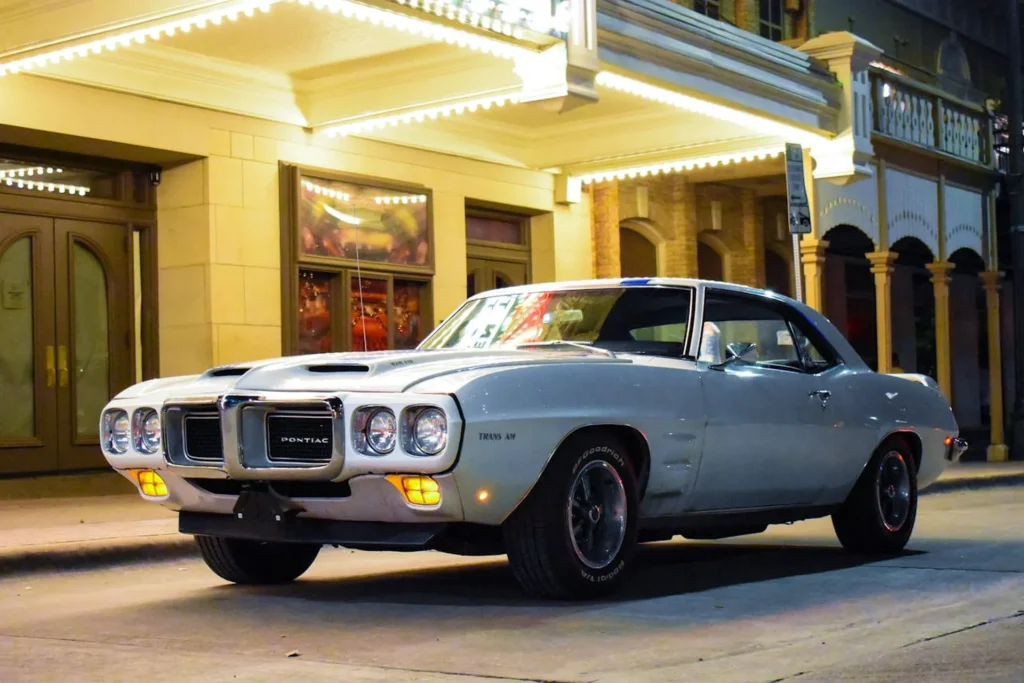 Na zdjęciu widzimy Pontiac Firebird Trans-Am zaparkowany przed eleganckim budynkiem z neonowymi światłami. Samochód jest w doskonałym stanie, z charakterystycznymi dwoma wlotami powietrza na masce i klasycznym designem lat 70-tych. Otoczenie sugeruje, że zdjęcie zostało zrobione w nocy, co podkreśla oświetlenie budynku w tle.