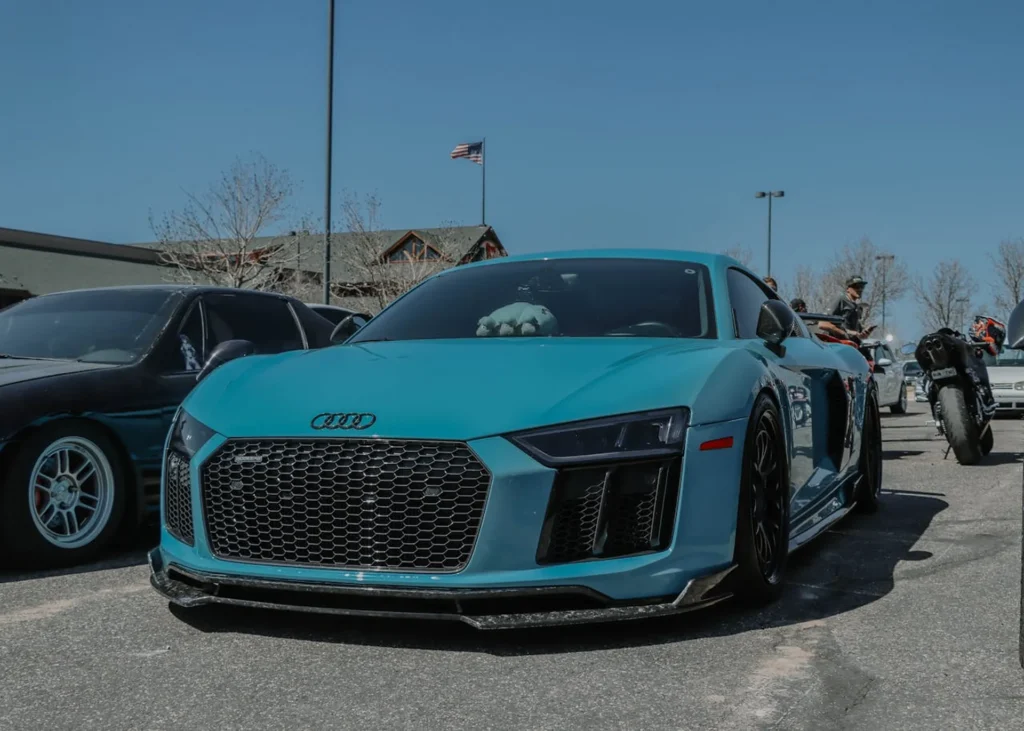 Niebieskie Audi R8 na zlocie samochodowym, z agresywnym grillem i karbonowymi detalami, w otoczeniu innych sportowych aut i motocykli.