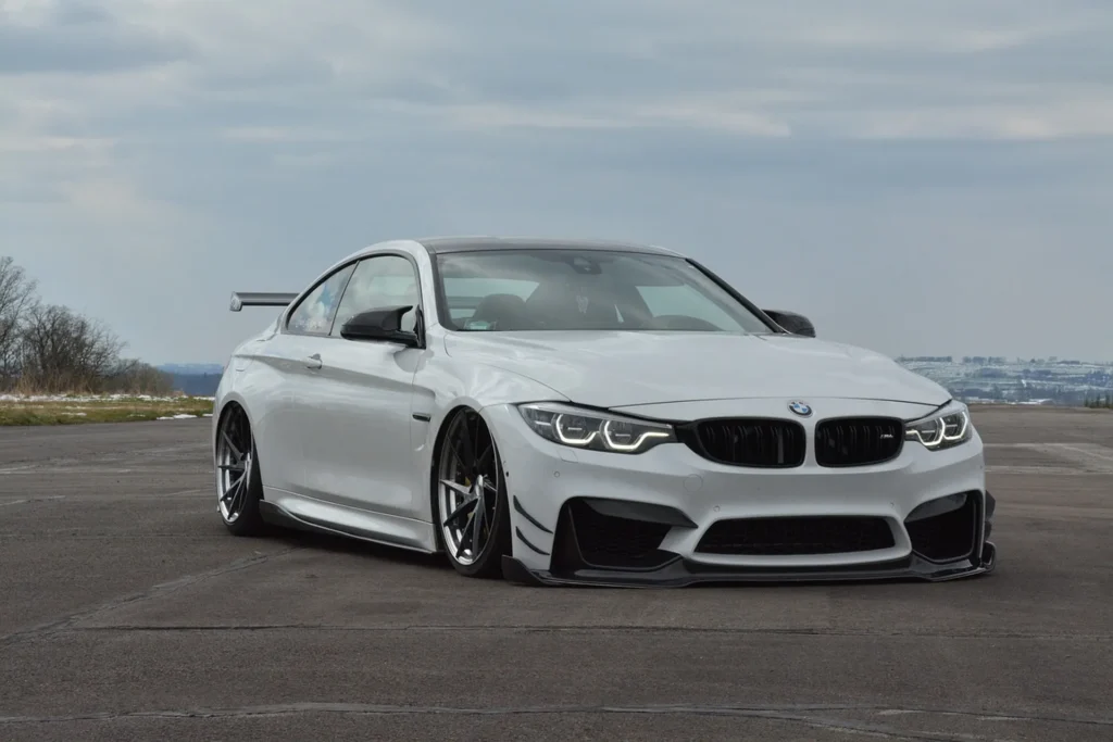 Białe BMW M4 z aerodynamicznym body kitem i niskim zawieszeniem, zaparkowane na otwartej przestrzeni pod pochmurnym niebem.