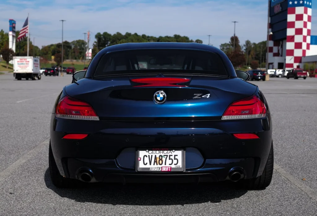 Tył granatowego BMW Z4 z tablicą rejestracyjną z Georgii, zaparkowanego na tle toru wyścigowego i amerykańskich flag.