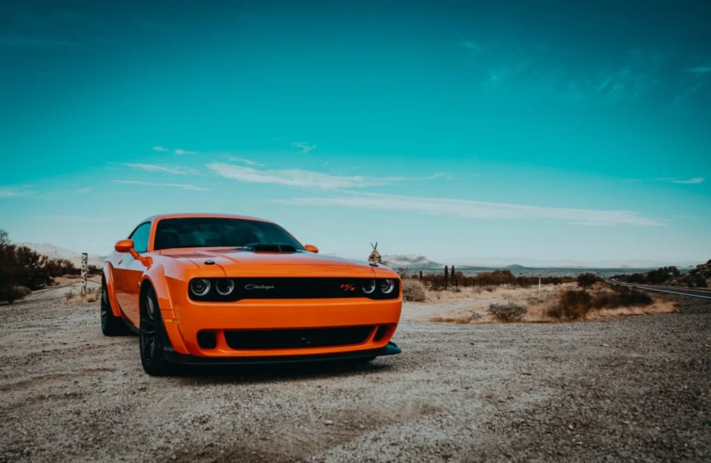 Pomarańczowy Dodge Challenger zaparkowany na pustynnym terenie pod jasnym, błękitnym niebem, z muskularnym nadwoziem i sportową sylwetką.