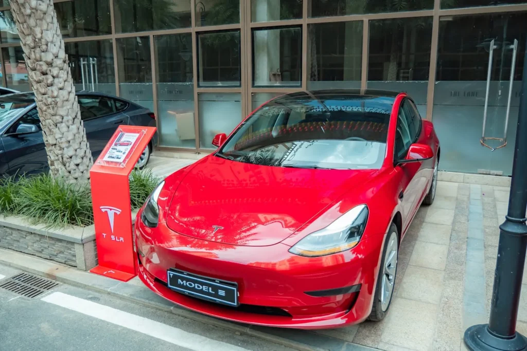Czerwona Tesla Model 3 zaparkowana przy stacji ładowania, z nowoczesnym designem i refleksami światła na karoserii.