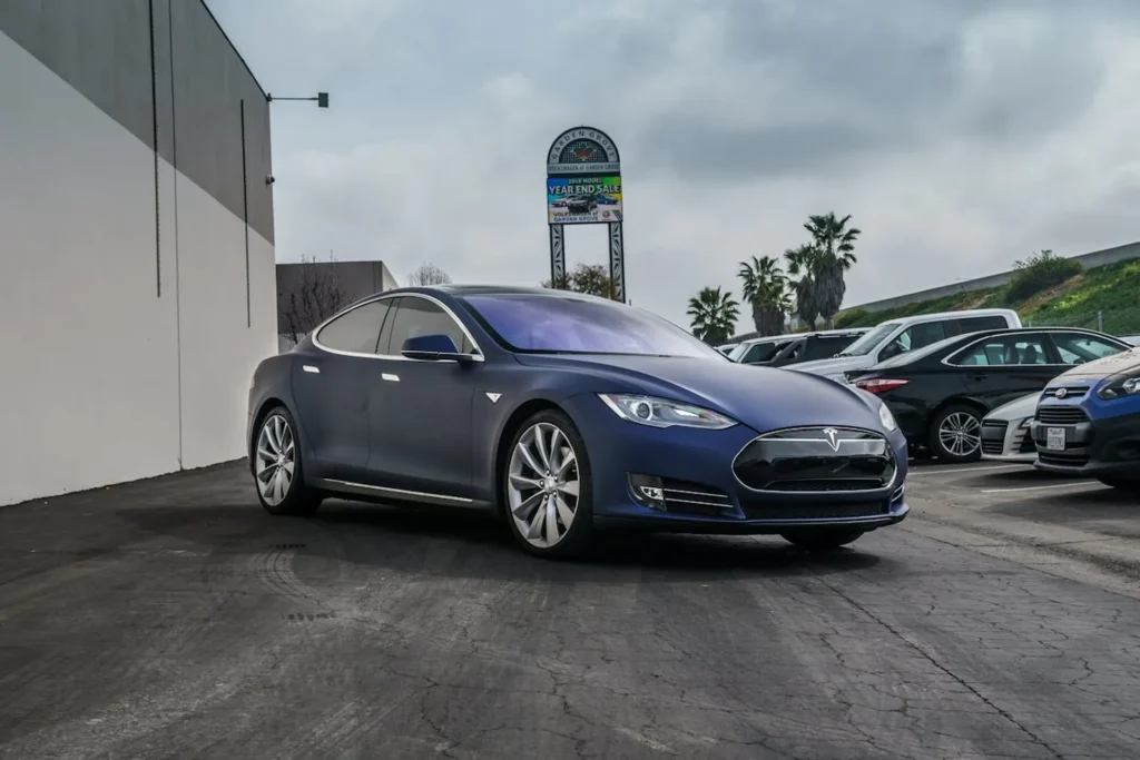 Granatowa Tesla Model S zaparkowana na parkingu obok innych samochodów, z elegancką sylwetką i charakterystycznym grillem.