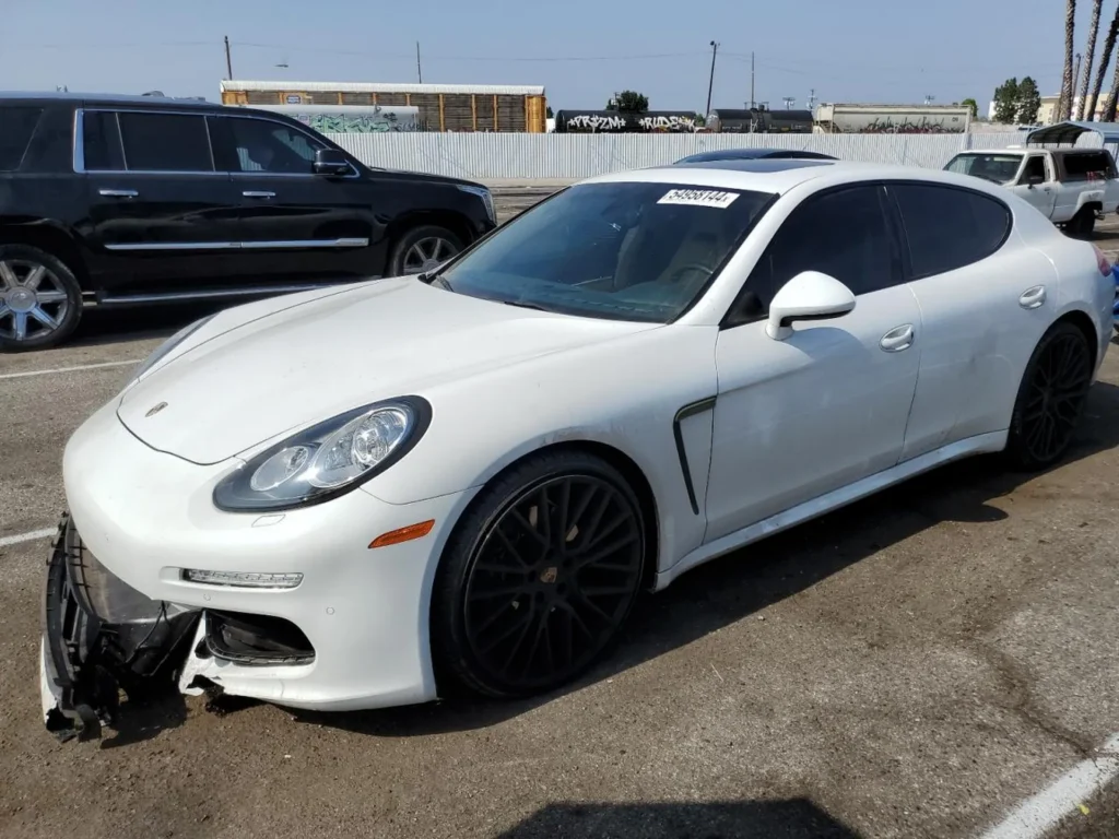 Biały Porsche Panamera – ujęcie z przodu lewego boku; zdemontowany/urwany dolny zderzak po lewej, czarne felgi, przyciemnione szyby.
