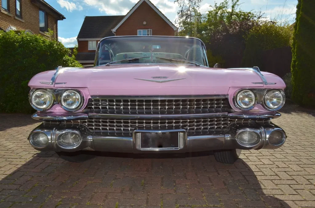Różowy Cadillac z lat 50. zaparkowany na podjeździe przed domem, sfotografowany od frontu, z charakterystycznym chromowanym grillem i podwójnymi reflektorami.