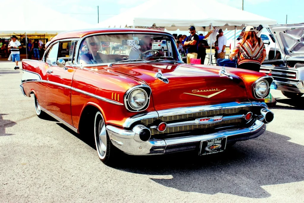 Czerwony Chevrolet Bel Air z 1957 roku, lśniący chromowanymi detalami, zaparkowany na zlocie klasycznych samochodów, z ludźmi i stoiskami w tle.