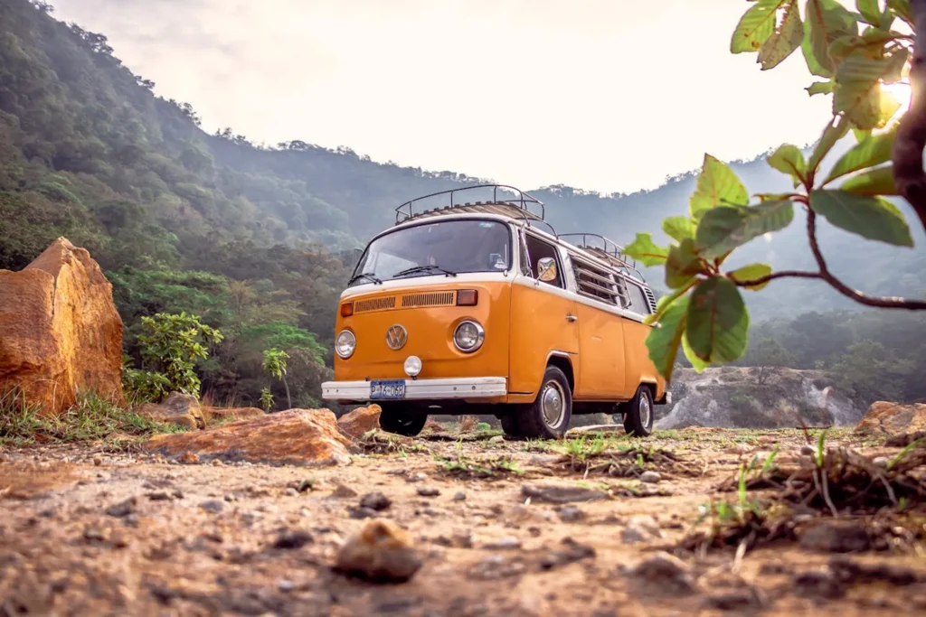 Pomarańczowy Volkswagen Transporter T2 zaparkowany na kamienistej drodze wśród górskiej roślinności, z bagażnikiem dachowym i klimatem podróżniczym.