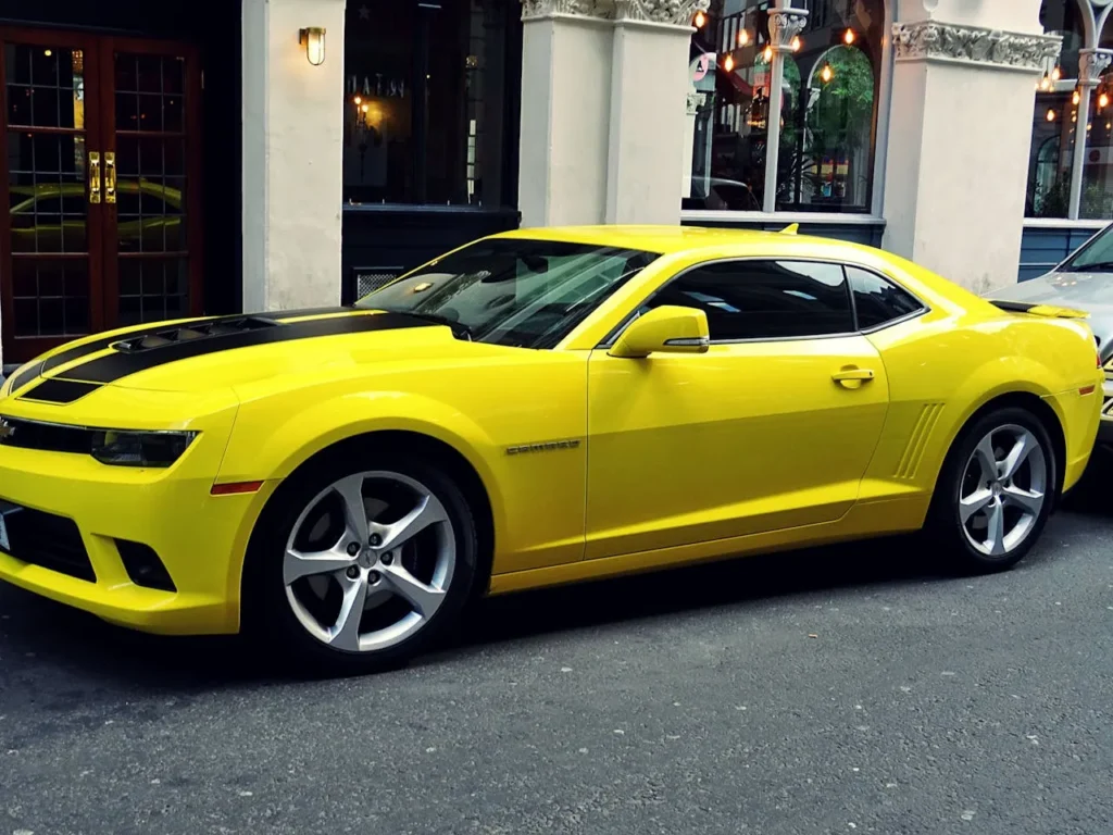 Żółty Chevrolet Camaro z czarnymi pasami na masce, zaparkowany na ulicy przed stylową restauracją, z nowoczesnym designem i sportowym charakterem.