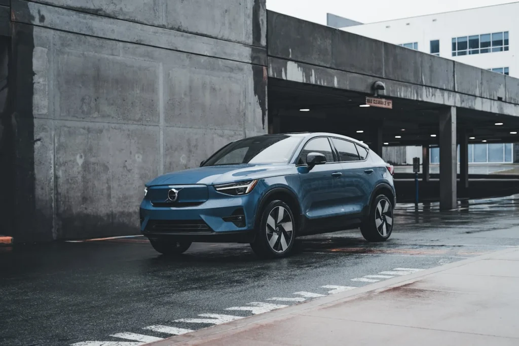 Niebieskie Volvo C40 Recharge zaparkowane na mokrej nawierzchni przy betonowym parkingu, z nowoczesnym designem i elektrycznym napędem.