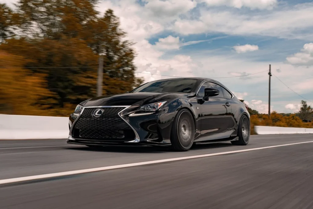 Czarny Lexus RC F Sport w ruchu na tle jesiennych drzew – sportowy coupe z charakterystycznym grillem w kształcie klepsydry.