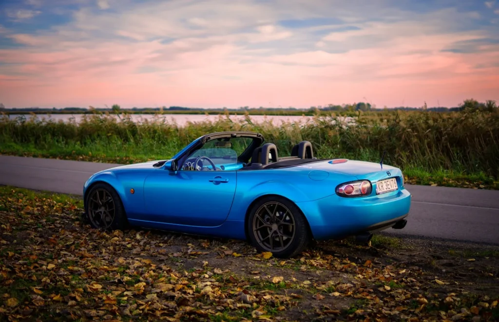 Niebieska Mazda MX-5 Miata z otwartym dachem zaparkowana nad rzeką w jesiennym krajobrazie