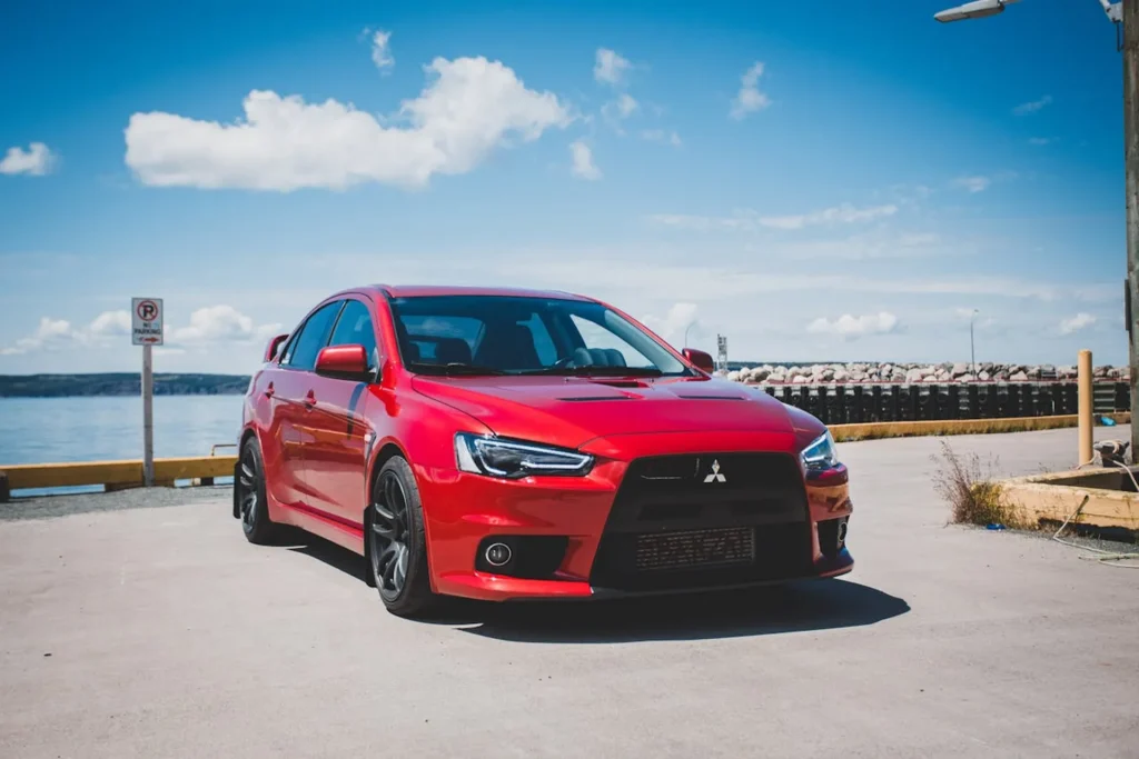 Czerwone Mitsubishi Lancer Evolution zaparkowane nad oceanem w słoneczny dzień, sportowa sylwetka i spoiler