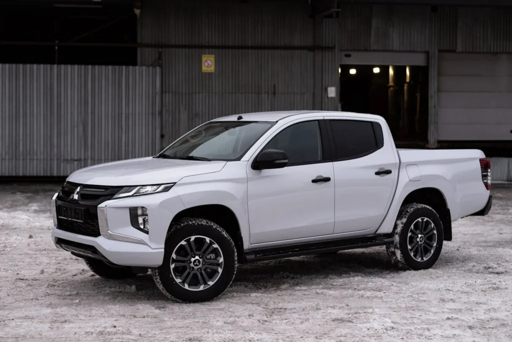 Biały pickup Mitsubishi L200 (Triton) zaparkowany na śniegu przed przemysłową halą