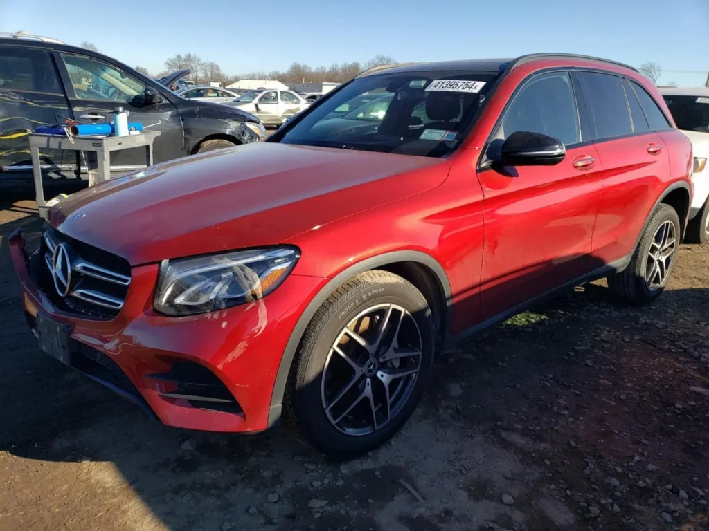 Czerwony Mercedes-Benz GLC 300 z lewego przodu na placu; felgi 5-ramienne, przyciemnione szyby, zarysowania na lewym zderzaku/reflektorze.