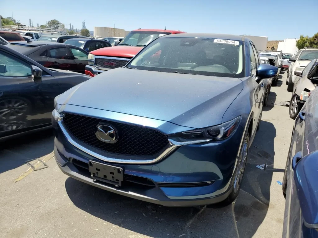 Niebieska Mazda CX-5 z przodu po lewej, zakurzona, bez tablic, stoi ciasno na parkingu aukcyjnym.