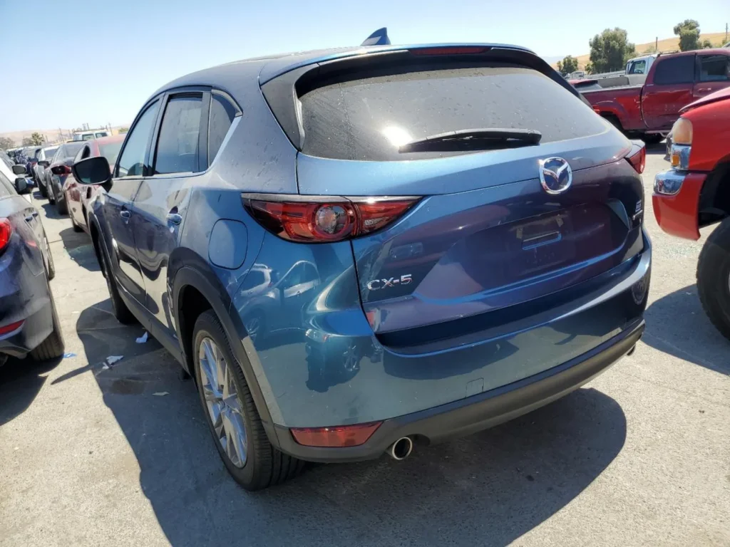 Tył i lewy bok niebieskiej Mazdy CX-5, zakurzone szyby, parking z autami w tle.
