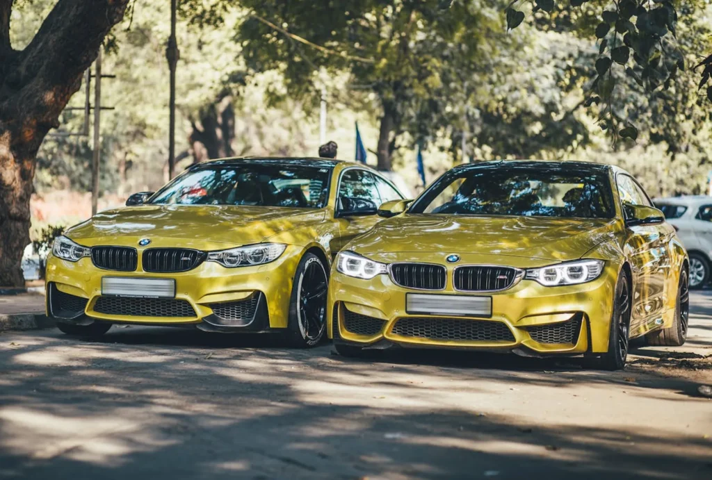 Dwa złote BMW M4 z USA zaparkowane obok siebie pod drzewami, widok z przodu