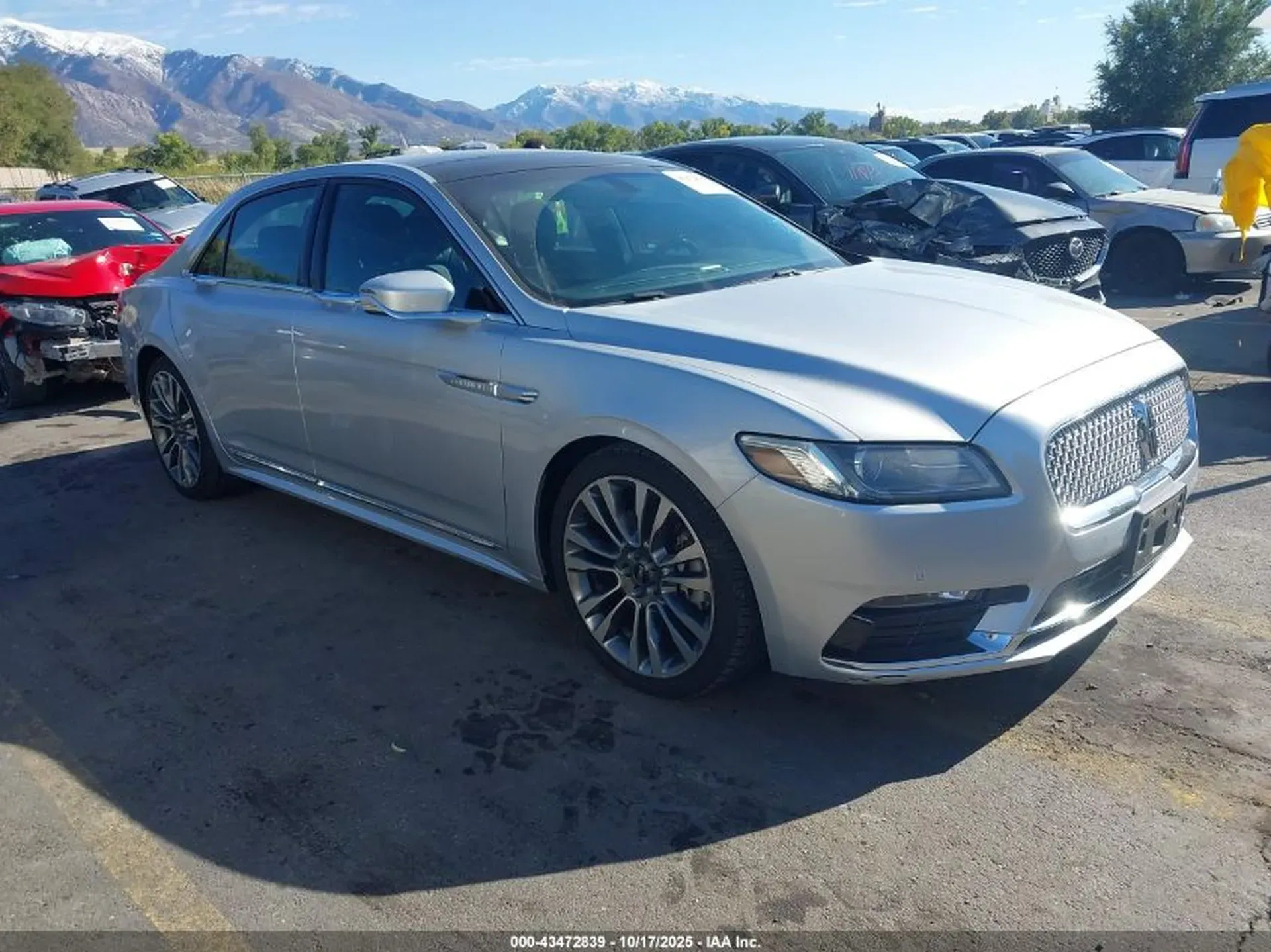 Srebrny Lincoln Continental z przodu z lewej, duży grill i góry w tle, parking aukcyjny.
