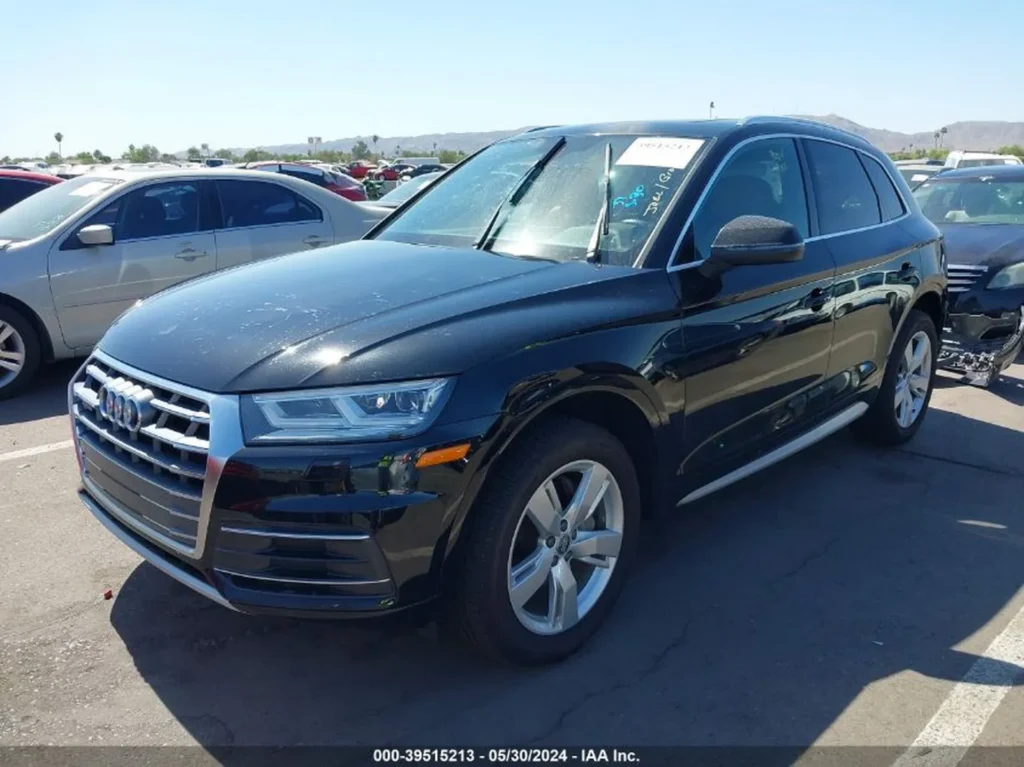 Audi Q5 z przodu z prawej, wycieraczki uniesione, auta w tle na placu.