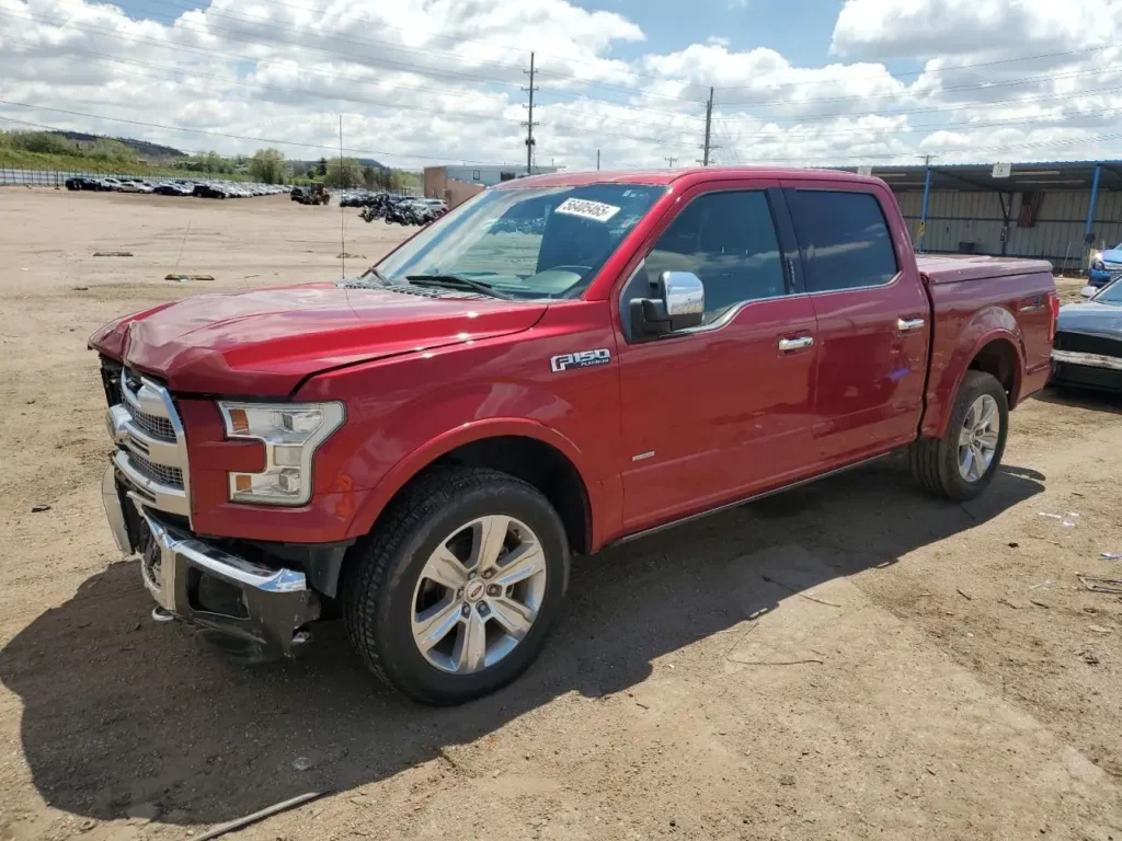 Czerwony Ford F-150 z podwójną kabiną i uszkodzonym przodem, stojący na piaszczystym placu.